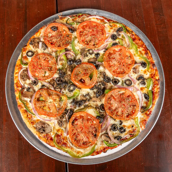 A whole vegetarian pizza with tomatoes, olives, mushrooms, onions, and green peppers on a metal pan.