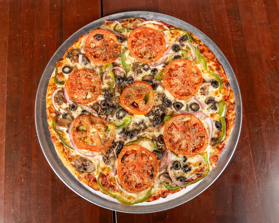 A whole vegetarian pizza with tomatoes, olives, onions, mushrooms, and green peppers on a metal tray.
